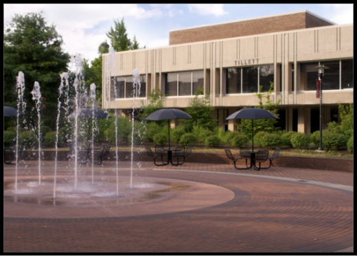 Image of the back of the Tillet Hall building with fountains and black picnic tables and umbrellas on brick outside.
