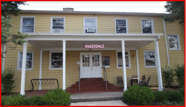 Image of the exterior of the Makerspace building. It is yellow with white posts and has a small wooden sign that has Makerspace written on it.