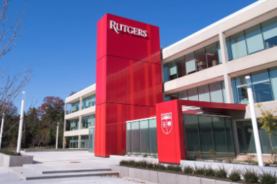 Image of the external of a building with a red tower in front with Rutgers on it and a smaller red part of the building with a shield on it. The building they are attached to is white with lots of windows.